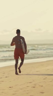 Kolunun altında sörf tahtasıyla sahilde koşan bir sörfçü.