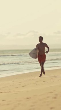 Gün batımında sörf tahtasıyla sahilde koşan genç sörfçü.