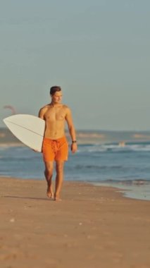 Genç sörfçü sörf tahtasıyla sahilde yürüyor.