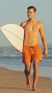 Genç sörfçü sörf tahtasıyla sahilde yürüyor.