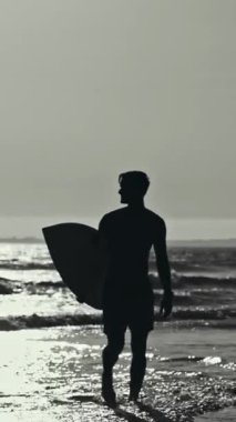 Gün batımında sörf tahtasıyla okyanusa yürüyen bir sörfçünün silüeti.