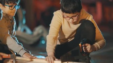 Bir atölyede birlikte çalışan iki genç çocuk, biri el testeresiyle tahta testereyi kesiyor.