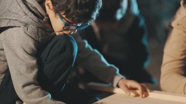 Gözlüklü çocuk marangozluk öğreniyor ve diğer çocuklarla birlikte atölyede tahta bir kalasa dokunuyor.