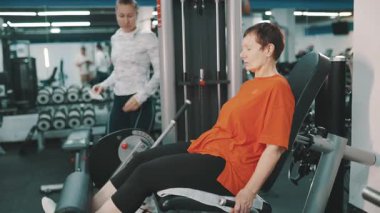 Yaşlı bir kadın spor salonunda beden eğitimi hocasıyla bacaklarını çalıştırıyor.