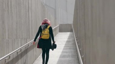 Sarı sırt çantalı genç bir kadın şehir ortamında modern beton bir rampada yürüyor.