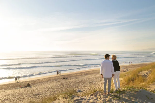 Kıdemli çift gün batımında okyanusa bakan kumulda sevgiyle kucaklaşıyorlar.
