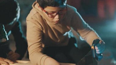 Gözlük takan, tahta testereyle tahta keserek marangozluk becerisi öğrenen odaklanmış bir çocuk.