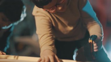 Gözlüklü genç bir çocuk atölyede odun keserek marangozluk becerileri öğreniyor.