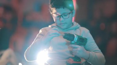 Gözlüklü konsantre çocuk atölyede kablosuz matkapla odun parçaları birleştiriyor.