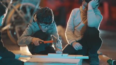 Gözlüklü genç, gece atölyesi sırasında tahta tahtaya çivi çakmayı öğreniyor.