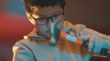 Yoğunlaşmış, gözlüklü, çekiçle tahtaya çivi çakan genç bir çocuk.