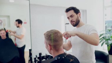 Genç bir erkek kuaför modern, parlak bir kuaförde müşterinin saçını buduyor ve tarıyor.
