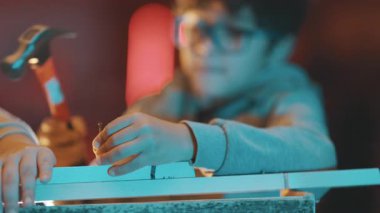 Gözlüklü yaratıcı çocuk ahşap işçiliği öğreniyor ve tahta çivi çakıyor.