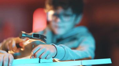 Gözlüklü yaratıcı çocuk marangozluk öğreniyor ve atölyede tahta çivi çakıyor.