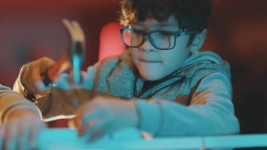 Gözlüklü küçük çocuk bir atölyede tahta bir çiviyi çekiçlerken konsantre oluyor.