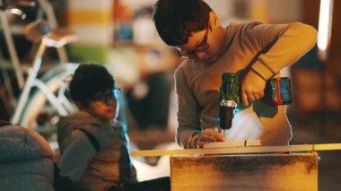 Arkadaşının atölyede izlediği gibi tahtada kablosuz elektrikli matkap kullanan odaklanmış bir çocuk.