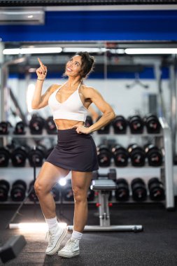 Zinde bir kadın gülümsüyor, parmağını yukarı kaldırıyor, modern bir spor salonunda fikir ya da yön gösteriyor.