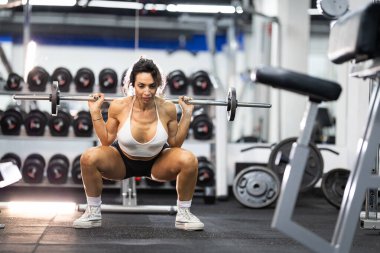 Atletik kadın halterle çömelmiş, spor salonunda antrenman yapıyor.