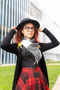 Üniversite kampüsünde ekose etek ve şapkayla şık şehir kıyafetleri içinde poz veren genç bir kadın.