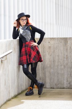 Beton duvara karşı poz veren şık bir kadın. Şapka, ekose etek ve çizme giyiyor.