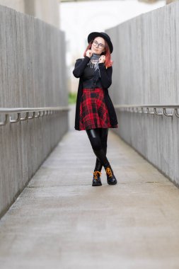 Ekose etekli, siyah şapkalı ve gözlüklü genç bir kadın şehir ortamında poz veriyor.