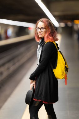 Metroda duran, arkasına bakan, sırt çantası takan ve şapkasını tutan genç bir kadın.