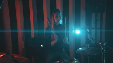 Genç bir kadın sinematik ışıklandırmalı bir müzik stüdyosunda enerjik bir şekilde davul çalıyor.