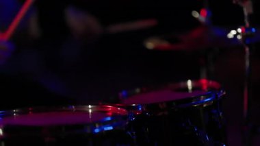 Müzisyen elleri kırmızı ve mavi ışıklarla karanlık bir sahnede davul çalıyor.