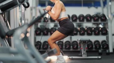 Atletik kadın modern bir spor salonunda kablo makinesiyle çömeliyor ve arka planda halter askısı var.