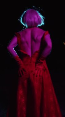 Siyah arka planda canlı ışıklar altında poz veren kırmızı bir gece elbisesi içinde zarif bir transseksüel.