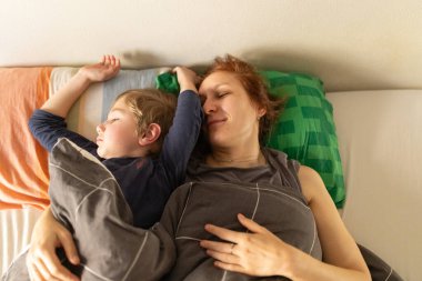 Anne ve oğlu battaniyenin altında birlikte uyuyorlar, aile bağlarını ve güvenliği resmediyorlar.