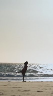 Gün batımında sahilde tek başına dans eden uzun kırmızı elbiseli zarif genç bir kadın. Orta çekim