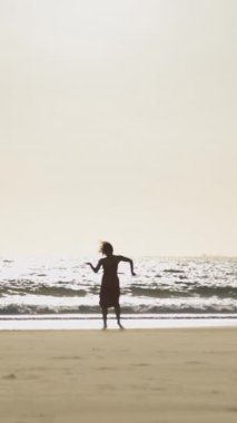 Gün batımında sahilde tek başına dans eden uzun elbiseli zarif genç bir kadın. Orta çekim