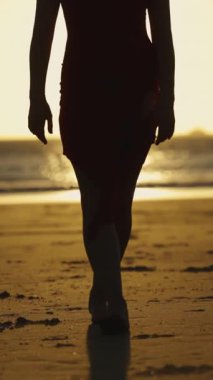 Gün batımında kumda çıplak ayakla yürüyen kırmızı elbiseli bir kadın. Orta çekim