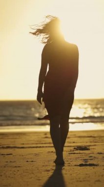 Gün batımında kumda çıplak ayakla yürüyen kırmızı elbiseli genç bir kadın. Orta çekim