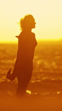 Parlak turuncu gün batımında, gece elbisesi giymiş bir kadın denizde yürüyor. Orta çekim