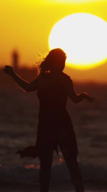 Parlak turuncu günbatımında, deniz kenarında dans eden genç bir kadının karanlık silueti. Orta çekim