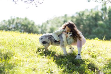 Genç bir kadın güneşli bir tepede çoban köpeğine sevgiyle sarılıyor.