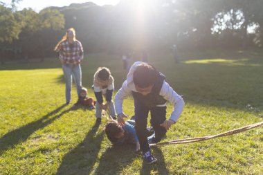Bir grup genç, parlak güneş ışığında yeşil çimlerin üzerinde iple aktif bir oyun oynuyor.