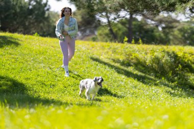 Genç bir kadın dışarıda antrenman yapıyor, güneşli bir günde köpeğiyle parkta koşuyor.
