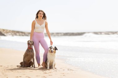 Okyanus dalgalarının olduğu kumlu bir sahilde çıplak ayaklı neşeli köpekleriyle duran bir kadın.