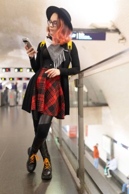 Platformda duran, korkuluklara yaslanan, toplu taşıma merkezinde akıllı telefon kullanan genç bir kadın.