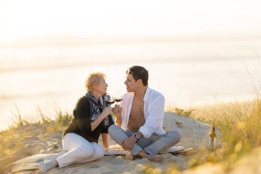 Kıdemli çift kumsalda altın saat günbatımında kırmızı şarap içiyorlar.