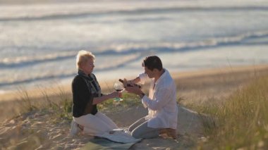 Zarif bir anne ve yetişkin oğul kumlu bir kumulda kırmızı şarapla romantik bir pikniğin tadını çıkarıyor.