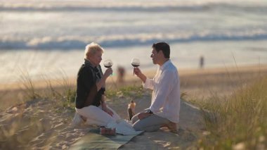 Yaşlı çift, kumlu bir kumulda romantik bir piknik sırasında kırmızı şarap içerken arkada okyanus vardı.
