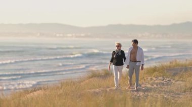 Mutlu olgun kadın ve genç adam gün batımında güzel bir sahil şeridinde el ele tutuşuyorlar.