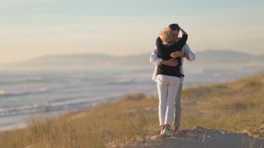 Güzel, olgun bir çift gün batımında arka planda okyanus olan kum tepeciğinde dans ediyor.