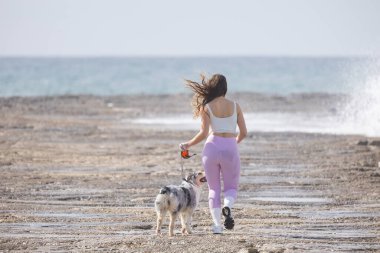 Kayalık bir sahil şeridinde tasmalı bir köpekle koşan bir kadın. Dalgalar kırılıyor.