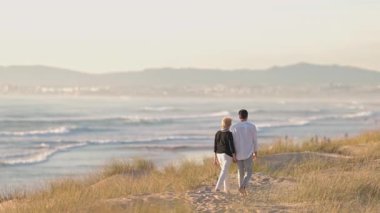Yaşlı çift el ele tutuşuyor ve kumulda yalın ayak yürüyor. Sarılmak ve denizi izlemek için duruyorlar.