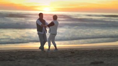 Mutlu, olgun bir çift, güzel bir günbatımında deniz kenarında el ele tutuşup dans ediyorlar.
